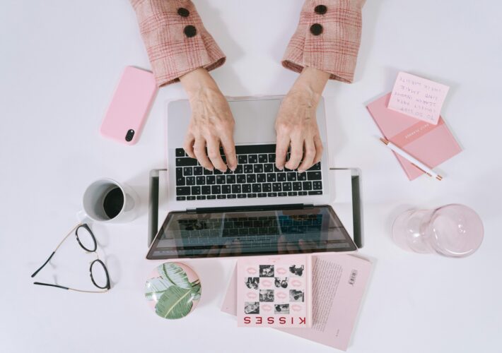 an image of a woman writing on her laptop on a messy pink desktop