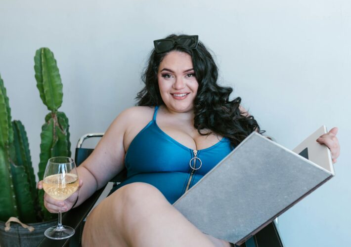 an image of a plus size woman in a swimsuit with a glass of wine in one hand and a large book in the other.