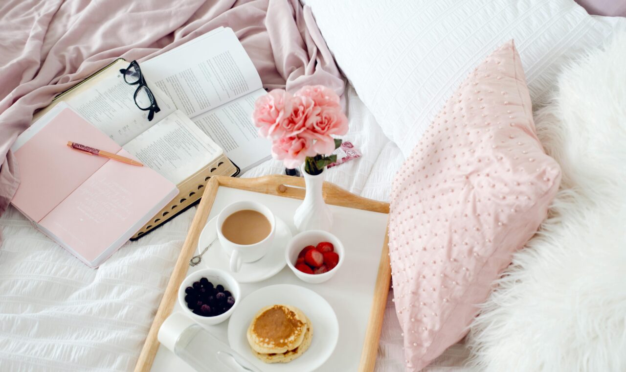 an image of cozy bed that has a breakfast tray full of food, and coffee. And next to it is a stack of books.