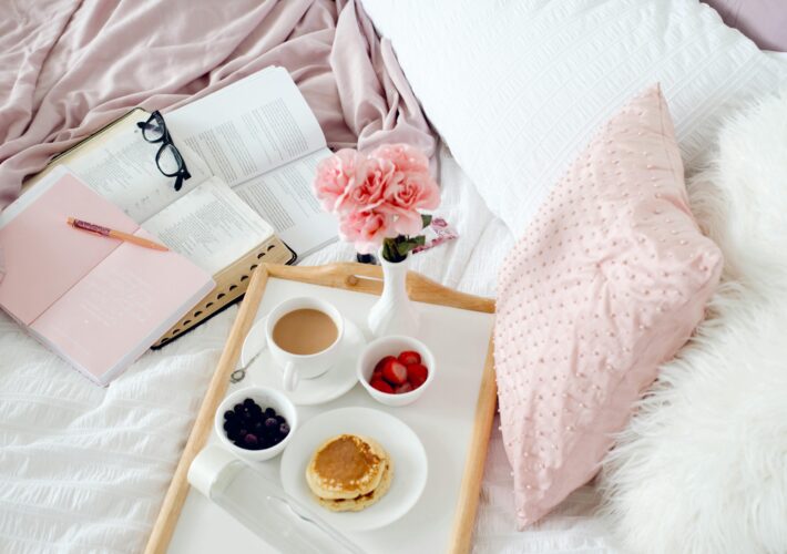 an image of cozy bed that has a breakfast tray full of food, and coffee. And next to it is a stack of books.