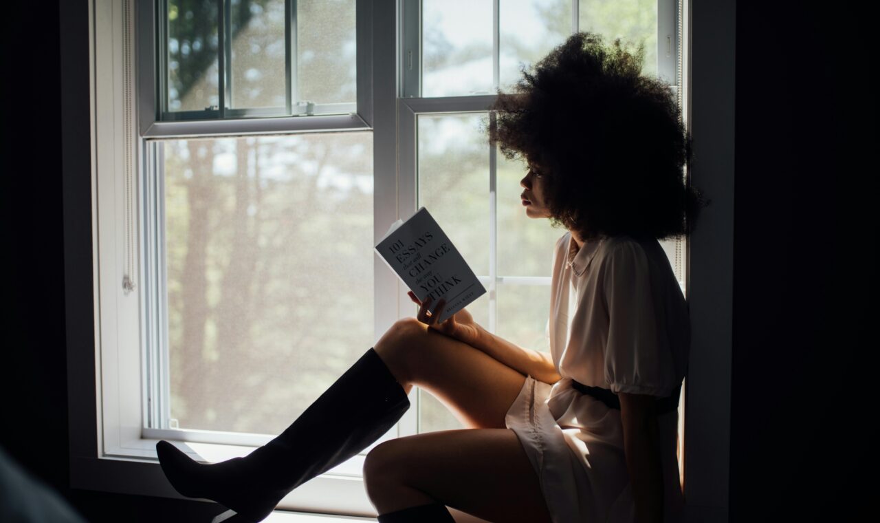 an image of a black woman with a large afro reading by the window.