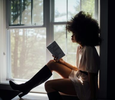 an image of a black woman with a large afro reading by the window.