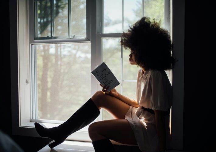 an image of a black woman with a large afro reading by the window.