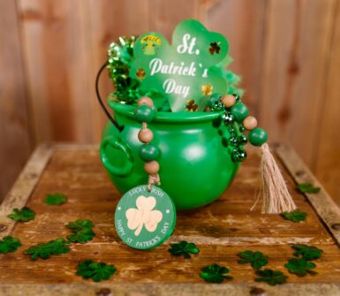 a green cauldron with a big green shamrock with some green beads stuck in it sitting on a table scattered with four leaf clovers