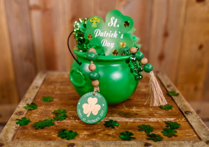 a green cauldron with a big green shamrock with some green beads stuck in it sitting on a table scattered with four leaf clovers