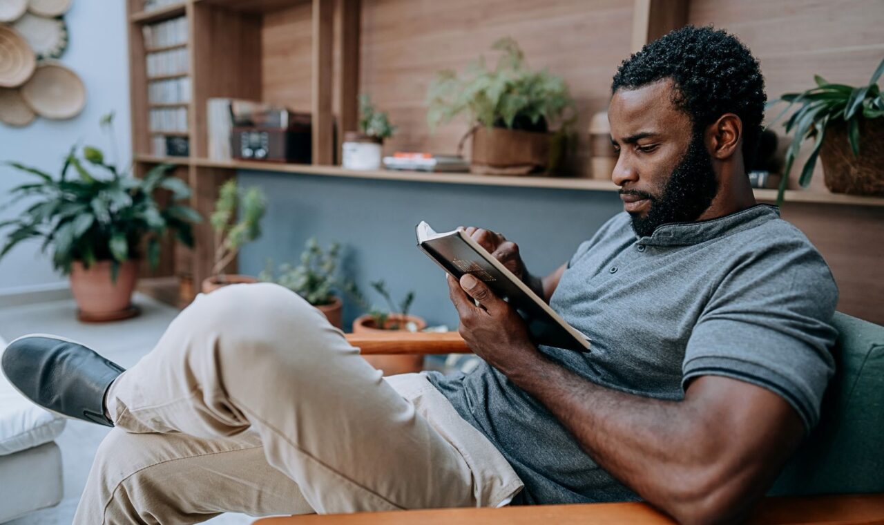 A man relaxes in a chair, reading a tablet in a cozy, plant-filled room. He appears focused, wearing a gray shirt and beige pants, surrounded by books and greenery.