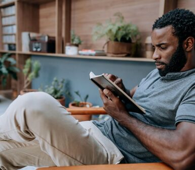 A man relaxes in a chair, reading a tablet in a cozy, plant-filled room. He appears focused, wearing a gray shirt and beige pants, surrounded by books and greenery.