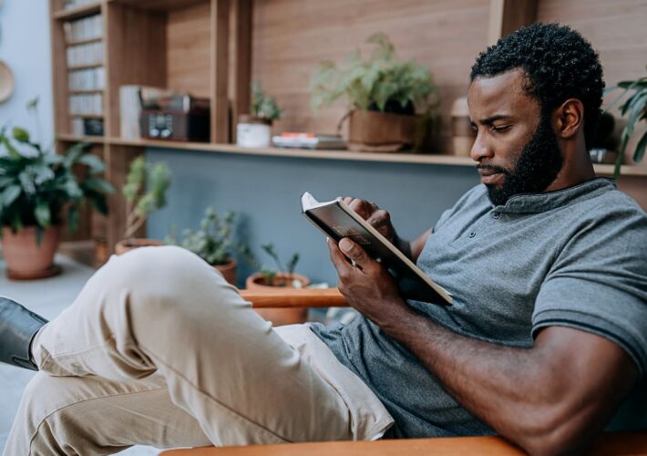 A man relaxes in a chair, reading a tablet in a cozy, plant-filled room. He appears focused, wearing a gray shirt and beige pants, surrounded by books and greenery.