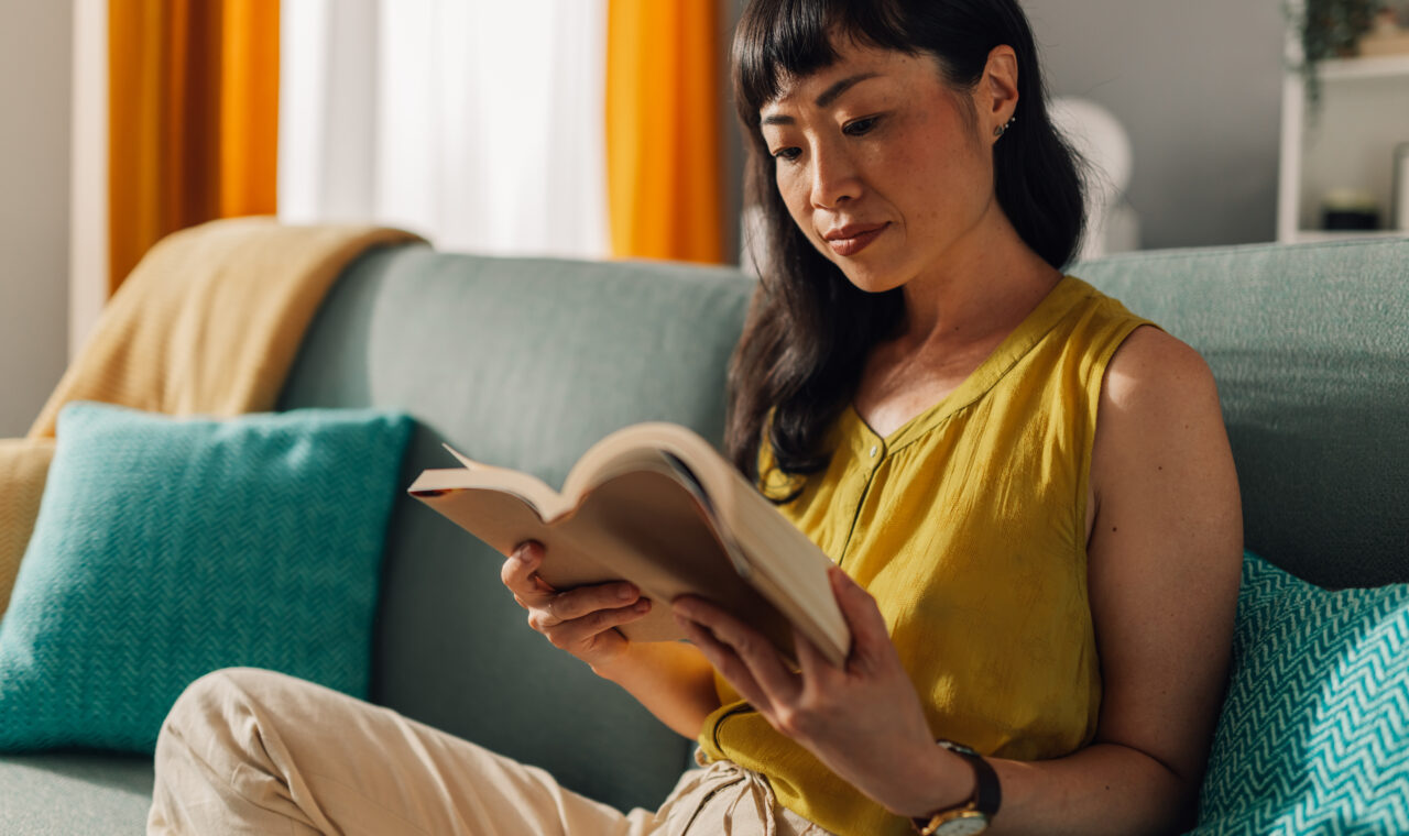 Asian woman in a yellow blouse and beige slacks sits on a teal couch, deeply focused on reading a book, surrounded by colorful cushions and a bright, inviting living room setting.