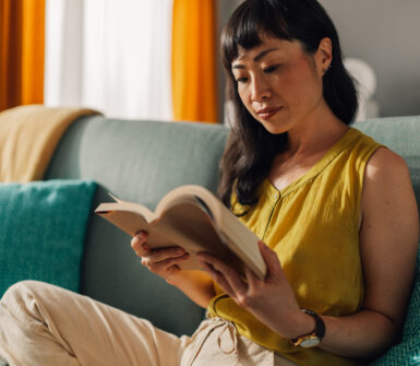 Asian woman in a yellow blouse and beige slacks sits on a teal couch, deeply focused on reading a book, surrounded by colorful cushions and a bright, inviting living room setting.