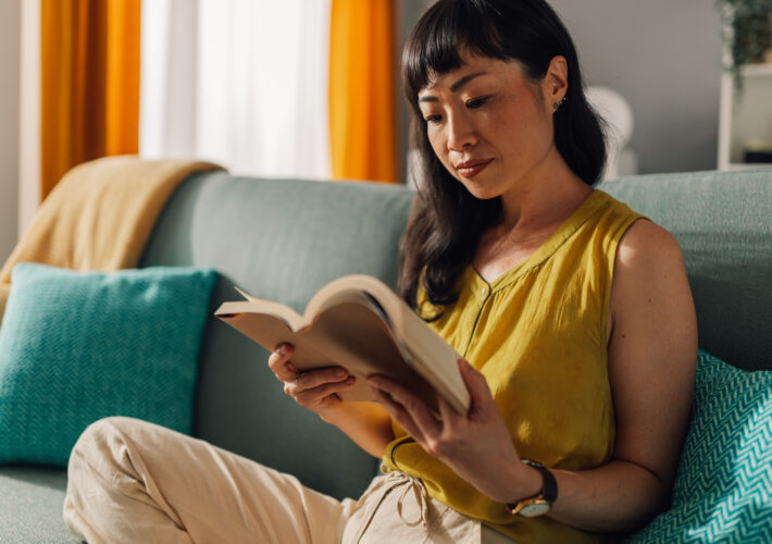 Asian woman in a yellow blouse and beige slacks sits on a teal couch, deeply focused on reading a book, surrounded by colorful cushions and a bright, inviting living room setting.