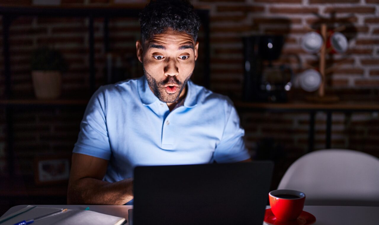 Hispanic man with beard using laptop at night afraid and shocked with surprise expression, fear and excited face.