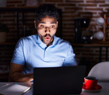 Hispanic man with beard using laptop at night afraid and shocked with surprise expression, fear and excited face.
