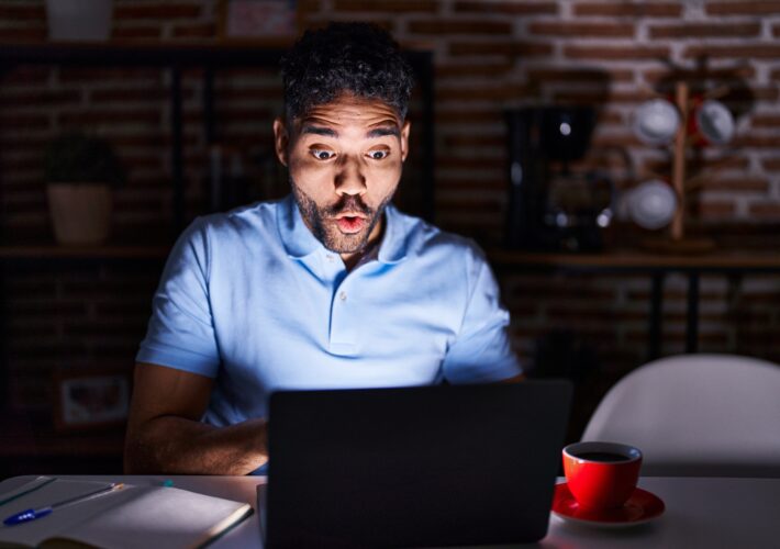 Hispanic man with beard using laptop at night afraid and shocked with surprise expression, fear and excited face.