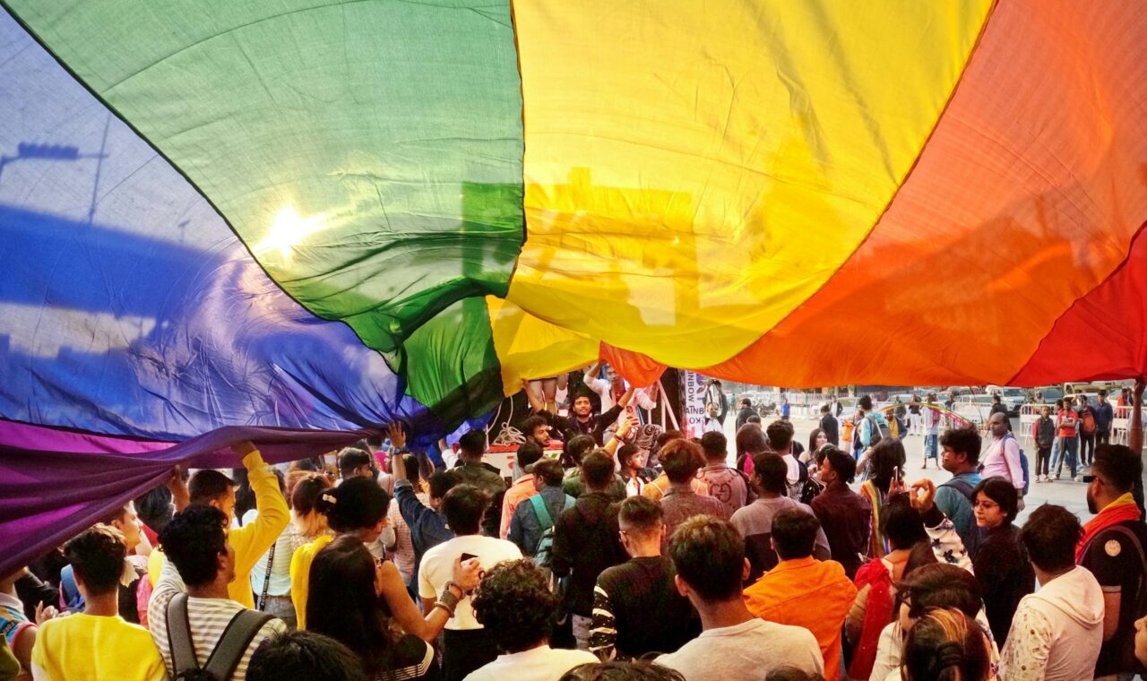 Joyful crowd celebrating Pride Parade under rainbow flag in Kolkata, India.