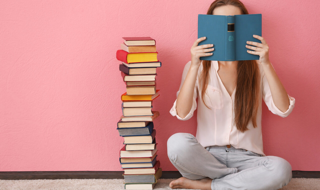 Woman sitting on a floor and holding book in front of face on pink background