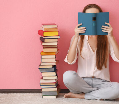 Woman sitting on a floor and holding book in front of face on pink background