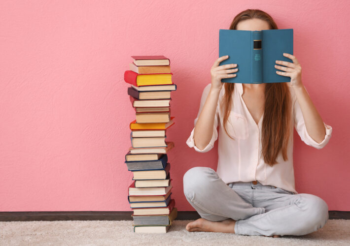 Woman sitting on a floor and holding book in front of face on pink background