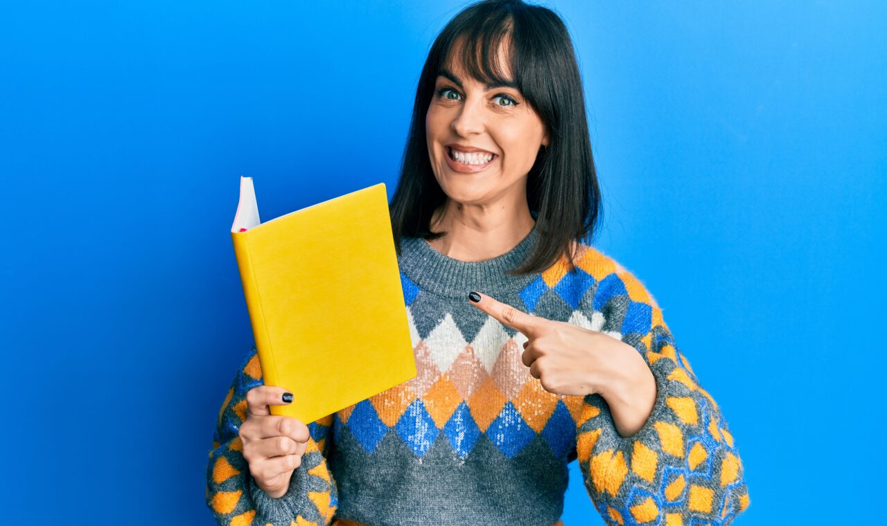 Young latina woman reading book smiling happy pointing with hand and finger