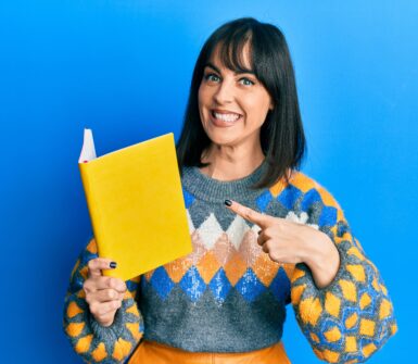 Young latina woman reading book smiling happy pointing with hand and finger