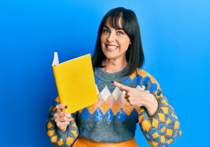 Young latina woman reading book smiling happy pointing with hand and finger