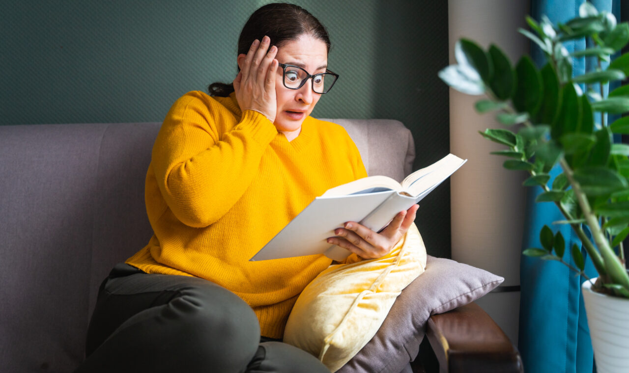 Frightened scared middle-aged woman in eyeglasses reading horror novel, thriller or scary stories while sitting on sofa at home, emotional shocked female with book in hands feeling terrified