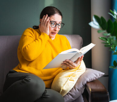 Frightened scared middle-aged woman in eyeglasses reading horror novel, thriller or scary stories while sitting on sofa at home, emotional shocked female with book in hands feeling terrified