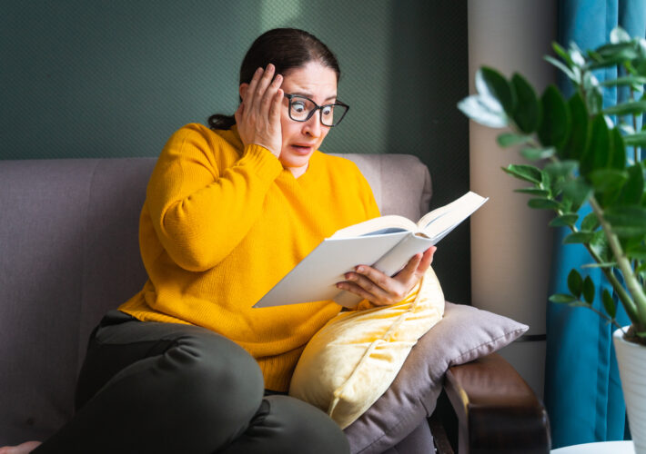 Frightened scared middle-aged woman in eyeglasses reading horror novel, thriller or scary stories while sitting on sofa at home, emotional shocked female with book in hands feeling terrified