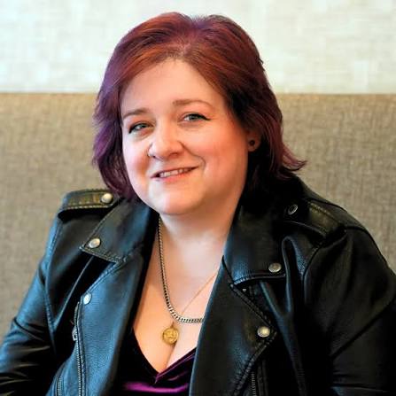 Portrait of a smiling woman with auburn hair wearing a black leather jacket and pendant necklace, seated indoors against a neutral background.