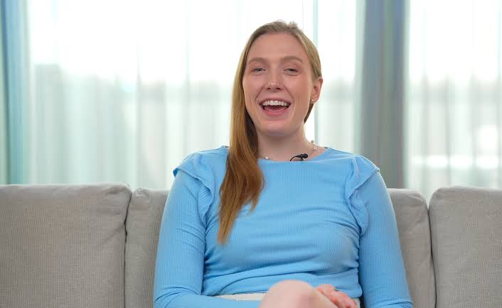 A picture of right wing influencer Pearl Davis.  She is a smiling woman in a light blue sweater sits on a gray sofa in a bright room, speaking to the camera. 